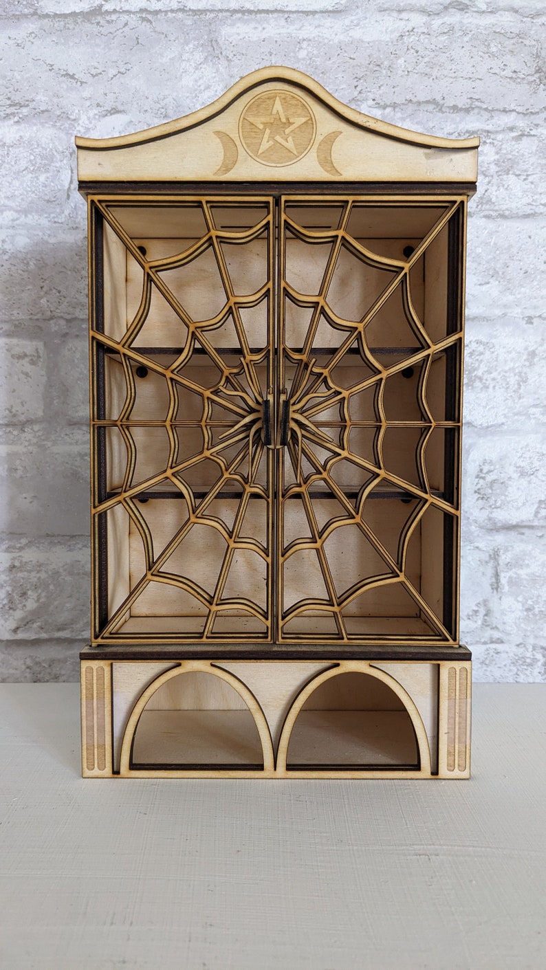Puede incluir: Un gabinete de madera con un dise&ntilde;o de telara&ntilde;a en las puertas. El gabinete tiene dos estantes y una base con dos aberturas arqueadas. La parte superior del gabinete tiene un pentagrama y un dise&ntilde;o de luna creciente.
