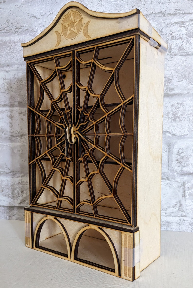 Puede incluir: Un gabinete de madera con un dise&ntilde;o de telara&ntilde;a decorativo y una luna creciente y un pentagrama en la parte superior. El gabinete tiene dos puertas y un estante debajo.