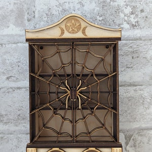 May include: A wooden cabinet with a spiderweb design on the doors. The cabinet has a shelf inside and a decorative top with a pentagram and crescent moons. The cabinet is painted in a light wood color with brown accents.