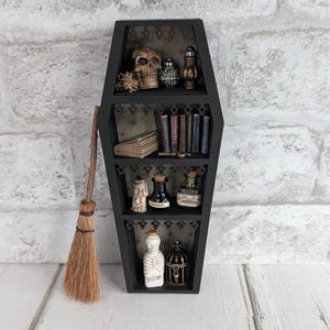 Pode incluir: Uma prateleira preta em forma de caixão com várias prateleiras, decorada com itens em miniatura com tema de Halloween. As prateleiras contêm uma caveira, livros, frascos de poções e uma estatueta de esqueleto. Uma pequena vassoura está encostada na prateleira.
