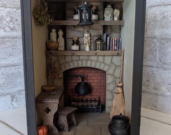 May include: Miniature witch's cottage diorama with a brick fireplace, shelves with potion bottles, books, and a hanging lantern. Includes a cauldron, broom, and pumpkins. The diorama is enclosed in a black frame.