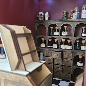 May include: A miniature apothecary shop with a wooden countertop, shelves, and drawers. The shelves are filled with small glass bottles with cork tops and labels. A skull and a candle are on the top shelf. The shop has a black and white checkered floor.