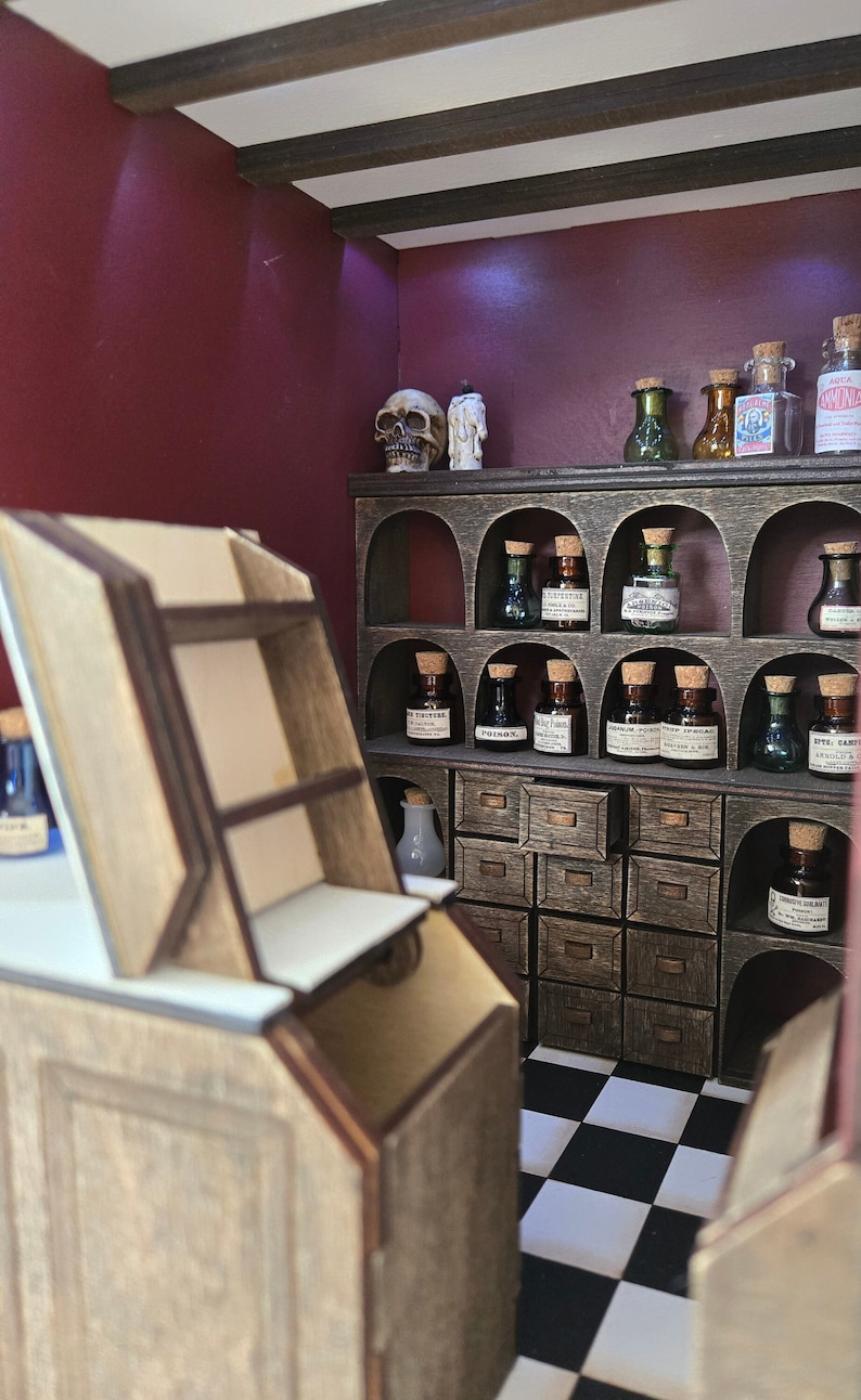 May include: A miniature apothecary shop with a wooden countertop, shelves, and drawers. The shelves are filled with small glass bottles with cork tops and labels. A skull and a candle are on the top shelf. The shop has a black and white checkered floor.