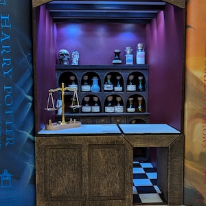 May include: A miniature apothecary shop with a wooden frame and shelves filled with glass bottles. The shop has a purple interior with a scale and a skull on the counter. The sign above the shop reads "Apothecary".