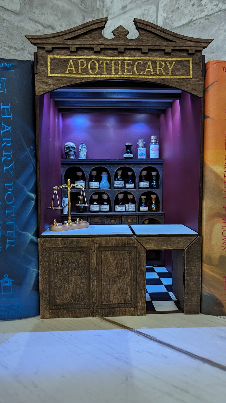 May include: A miniature apothecary shop with a wooden frame and shelves filled with glass bottles. The shop has a purple interior with a scale and a skull on the counter. The sign above the shop reads "Apothecary".