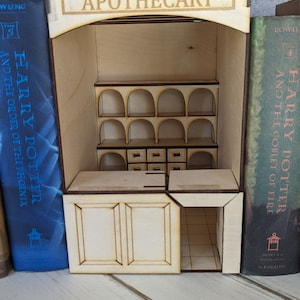 May include: A wooden apothecary shop model built from two Harry Potter book covers. The shop has a sign that reads "Apothecary" and features shelves with arched openings and drawers. The shop is open and the interior is visible.