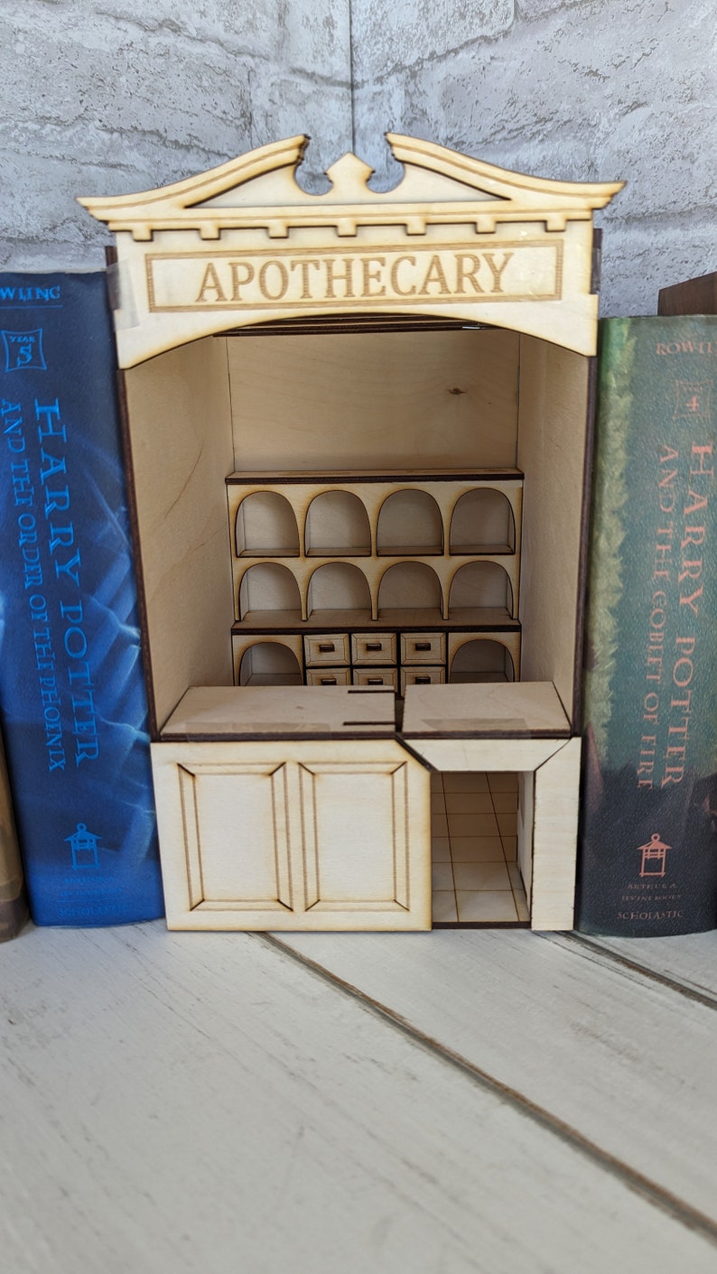 May include: A wooden apothecary shop model built from two Harry Potter book covers. The shop has a sign that reads "Apothecary" and features shelves with arched openings and drawers. The shop is open and the interior is visible.