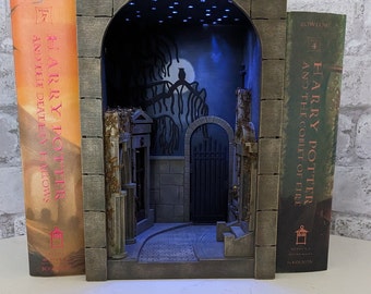 May include: A bookend shaped like a cemetery archway with a lit-up scene inside. The bookend is made of brown wood and features a small owl perched on a branch. The scene inside the archway is a dark, shadowy cemetery with a moon and stars in the sky. The bookend is flanked by two Harry Potter books, "Harry Potter and the Deathly Hallows" and "Harry Potter and the Goblet of Fire".