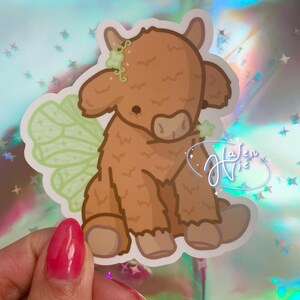 May include: A brown and white cartoon highland cow with green fairy wings. The cow has a flower crown on its head and is sitting down.