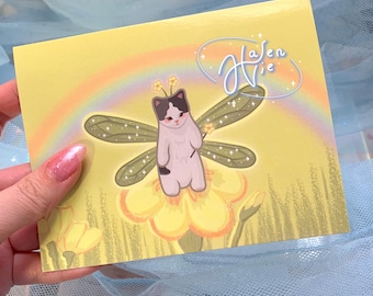 Puede incluir: Una tarjeta amarilla con un arcoíris y un gato blanco con manchas negras vestido de hada con alas verdes y una varita mágica. La tarjeta tiene el texto "Hasen Vie" en cursiva.