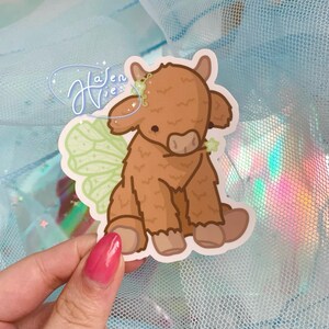 May include: A brown and white Highland cow with green fairy wings. The cow is sitting down and has a cute expression on its face.