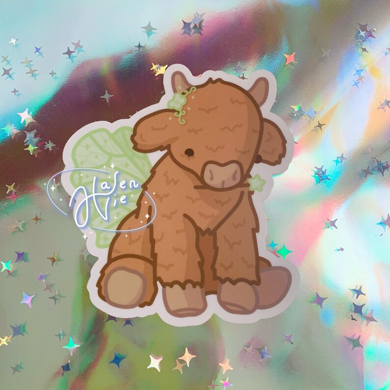 May include: A brown and white fuzzy cow with green fairy wings. The cow is sitting down with its head tilted down. The wings are made of leaves and have a green and white color scheme.