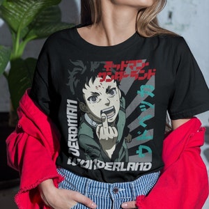 Peut inclure: T-shirt noir avec un imprimé graphique d'un personnage d'anime faisant un doigt d'honneur. Le personnage est vert et a une cicatrice rouge sur le front. Le texte "WONDERLAND" est imprimé sous le personnage. Le texte "JERKMAN" est imprimé à gauche du personnage. Le texte est en japonais.