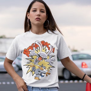 Può includere: T-shirt bianca con una grafica a cartoni animati di un coniglio e di un roditore in azione, con la parola "BOOM!" in rosso. La maglietta è indossata con jeans azzurri.