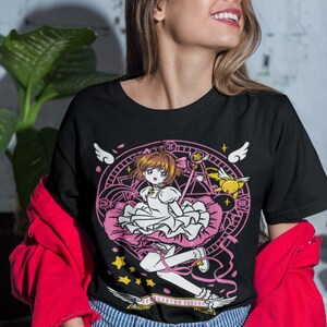 May include: Black t-shirt with a graphic print of a cartoon girl with pink hair and a pink dress holding a wand. The girl is surrounded by a circle with stars and wings. The text "Cardcaptor Sakura" is printed below the graphic.