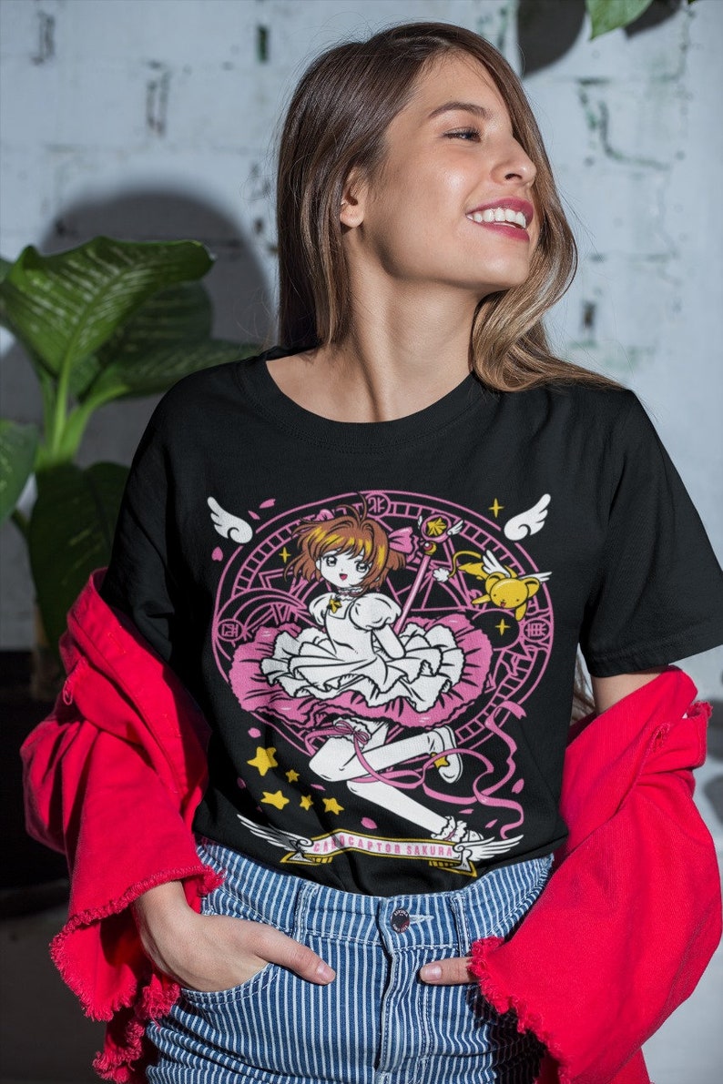 May include: Black t-shirt with a graphic print of a cartoon girl with pink hair and a pink dress holding a wand. The girl is surrounded by a circle with stars and wings. The text "Cardcaptor Sakura" is printed below the graphic.