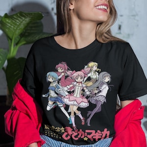 May include: Black t-shirt featuring a group of anime characters from the show "Puella Magi Madoka Magica" with the Japanese text "魔法少女まどか☆マギカ" (Mahou Shoujo Madoka Magica).