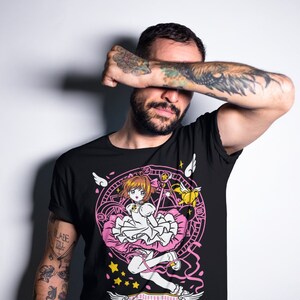 May include: Black t-shirt with a pink and white graphic of a girl with a pink dress and a wand, surrounded by a circle with stars and wings. The text "CARDCAPTOR SAKURA" is written on a banner below the graphic.