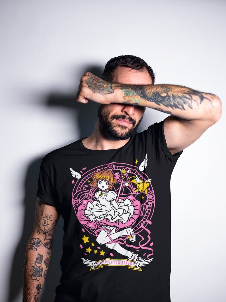 May include: Black t-shirt with a pink and white graphic of a girl with a pink dress and a wand, surrounded by a circle with stars and wings. The text "CARDCAPTOR SAKURA" is written on a banner below the graphic.