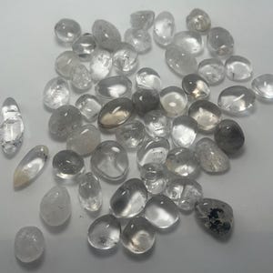 May include: A collection of clear quartz tumbled stones, some with a slight smoky or cloudy appearance. The stones are smooth and polished, with a variety of shapes and sizes.