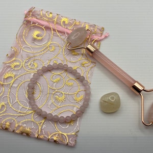 May include: A pink rose quartz facial roller, a pink rose quartz bracelet, and a pale green crystal stone on a pink and gold patterned fabric bag.