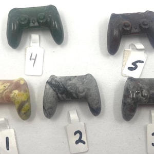 May include: A collection of carved stone game controller pendants in various colors. The pendants include green, brown, gray, and a mix of pink and yellow. Each pendant has a tag with a number.
