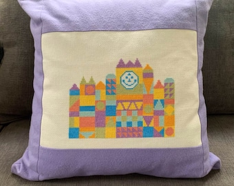 May include: A purple pillow with a white embroidered image of a colorful building with a smiling face on the clock tower.