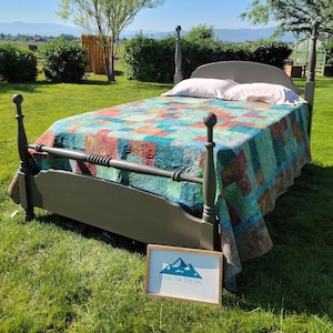 May include: A gray wooden bed frame with a colorful patchwork quilt. The quilt features squares of teal, red, and green. Two white pillows rest on the bed. A sign with the text "Under the Big Sky" is in front of the bed.