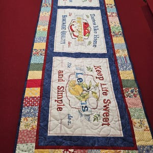 May include: A quilted table runner with three rectangular panels featuring fruit illustrations and text. The panels are bordered by blue fabric and surrounded by a patchwork design with floral and geometric patterns. The text includes "Peaches", "Sweetness", and "Lemons".