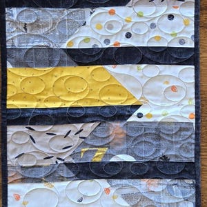 May include: A rectangular quilted table runner with a geometric design. The runner features alternating bands of yellow, black, and patterned fabrics. The quilting pattern includes circles and curved lines, creating a textured surface.
