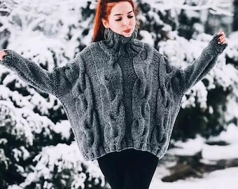 May include: A chunky knit sweater in a dark gray color featuring a prominent cable knit pattern throughout the body and sleeves. The garment has a high turtleneck collar, long sleeves with a relaxed fit, and a thick ribbed hem at the bottom edge.