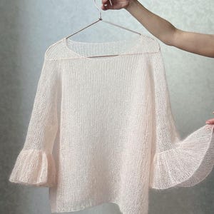 Könnte beinhalten: Ein zarter, blassrosa Strickpullover mit Glockenärmeln. Der Pullover ist aus einem leichten, luftigen Garn gefertigt und hat eine lockere, fließende Passform.
