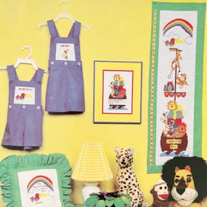 May include: A display of children's items and decor, including denim overalls with embroidered names, a framed cross-stitch, and a tall cross-stitch banner featuring animals. A stuffed leopard and a lion-faced plush are also present.