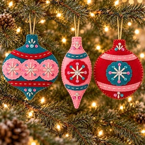 May include: Three felt Christmas ornaments hanging by gold strings. The left is a teal onion-shaped ornament with pink circular accents. The center is a pink teardrop-shaped ornament with a red center. The right is a red round ornament with a teal center.