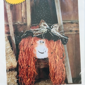 May include: A Halloween decoration featuring a scarecrow witch with orange raffia hair and a black hat with a polka dot pattern. The witch has a painted face with a smiling mouth and is adorned with small black spiders.