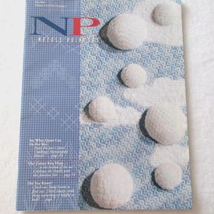 May include: A magazine cover titled "Needle Pointers" with a blue and white design. The cover features the letters "NP" in blue and red, with white, textured, circular shapes. Text on the left side includes article titles.