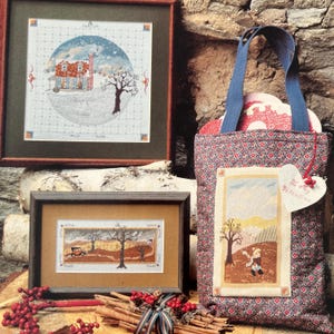 May include: A display of framed cross-stitch art and a tote bag. The art pieces depict winter and autumn scenes with houses, trees, and figures. The tote bag is patterned with a floral design and has a heart-shaped tag that says "To My Valentine."