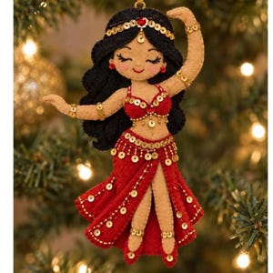 May include: A felt ornament depicting a dancer with long black hair, wearing a red two-piece outfit adorned with gold sequins and a red gem on the headband. The figure has tan skin, closed eyes, and arms raised in a graceful pose with gold accents.
