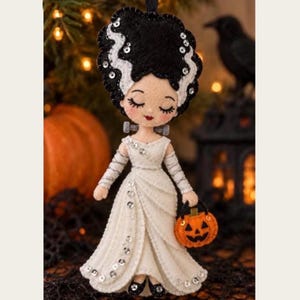 May include: A felt ornament depicting a character with tall black hair featuring white streaks and silver sequins. The figure wears a long white gown with silver sequin accents and holds a small orange felt pumpkin with a black jack-o'-lantern face.