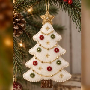 Peut inclure: Une décoration de sapin de Noël en feutrine crème avec une étoile dorée au sommet, un tronc marron et une guirlande de perles dorées. L'arbre est orné de petits cercles en feutrine rouges et verts et d'étoiles dorées brodées.