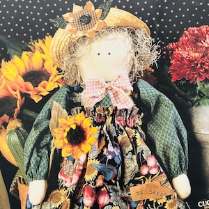 May include: A handmade doll with a straw hat adorned with a sunflower and a plaid bow. The doll wears a green quilted top and a floral print dress with a sunflower accent. The doll is surrounded by sunflowers and other fall harvest elements.