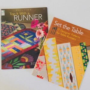 May include: Two books on a white surface. The book on the left is titled "Top It With a Runner" and features colorful quilt designs. The book on the right is titled "Set the Table" and showcases table runner patterns. A vase of pink flowers is visible.