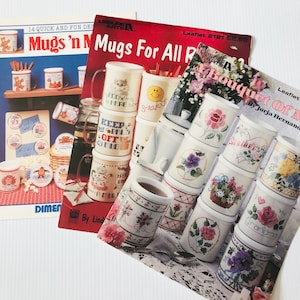 May include: A collection of vintage craft pattern books and mugs. The books feature cross-stitch designs and mug decorating ideas. The mugs have various designs, including floral patterns and text. The text on the books reads "Mugs 'n Mugs For All" and "A Bouquet Of Mugs".