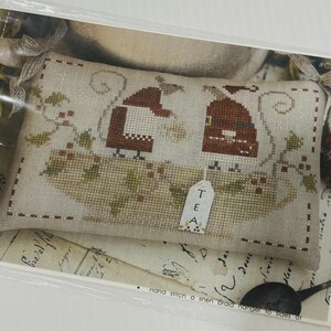 May include: A cross-stitched pillow with two figures in Santa hats, holding teacups, and the word "TEA" on a tag. The pillow is beige with brown and green accents, and a floral design.
