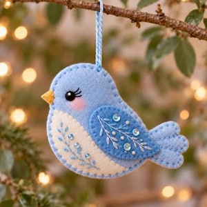 Peut inclure: Une décoration d'oiseau en feutrine bleu clair avec un poitrail blanc, un bec jaune et un œil noir à longs cils. L'aile présente des motifs de feuilles brodés en blanc et de petites paillettes bleues. Un cordon tressé bleu est fixé au sommet.