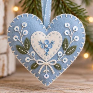 May include: A light blue felt heart ornament featuring a smaller white felt heart at the center. The design includes embroidered green leaves, white stitched edges, a white thread bow, and blue and white sequins arranged in floral patterns.