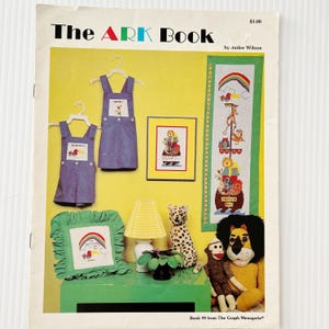 May include: The cover of "The ARK Book" features a yellow background with a variety of items. Two denim overalls hang on hangers. A framed picture and a long vertical banner are also displayed. A stuffed lion and monkey sit on a green surface.