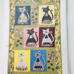 May include: A colorful cat-themed quilt featuring six cat portraits on a green background. Each cat is set against a different colored square, including blue, yellow, pink, and burgundy. The quilt is decorated with floral patterns and paw prints.