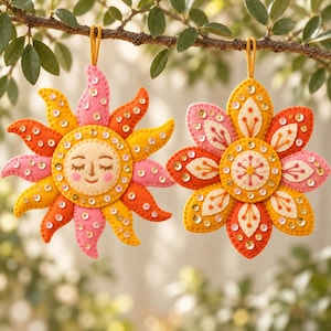 May include: Two felt hanging ornaments featuring a sun with a smiling face and a multi-petaled flower. Both ornaments are decorated with gold sequins and embroidered details in shades of pink, orange, yellow, and cream, suspended by gold cords.