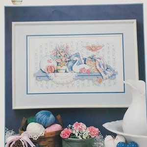 May include: A framed cross-stitch artwork depicts a shelf with a swan, flowers, and a basket. The color palette includes soft blues, pinks, and whites. A decorative angel and heart-shaped accents add to the vintage charm. The artwork is framed in a white mat and a blue frame.
